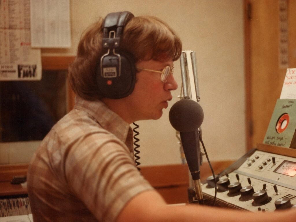 KCPR DJ wearing headphones and speaking into the studio microphone beside the control board